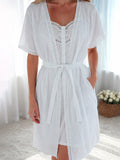 A woman wearing a white short-sleeve lace-trimmed robe with a tie waist, standing in a bright indoor setting with white curtains and a basket of flowers in the background.