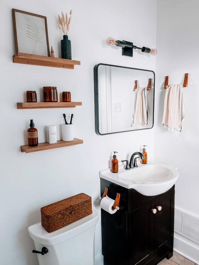 Modern bathroom featuring vegetable-tan leather towel racks and a matching vegetable-tan leather toilet paper holder with birch dowels, styled beside a dark vanity with amber soap bottles, floating wooden shelves, and a black framed mirror, showcasing Keyaiira’s handcrafted leather and wood Bathroom Bundle in a warm Scandinavian-inspired space.