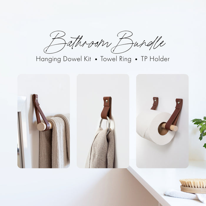Bathroom Bundle display showing three coordinated leather and wood accessories: a hanging dowel towel rack with warm leather straps holding a folded beige towel, a hand towel ring with leather and a metal ring supporting a textured beige towel, and a toilet paper holder using the same brown leather straps with a wooden dowel holding a roll of toilet paper. Minimalist white wall background with soft natural styling.