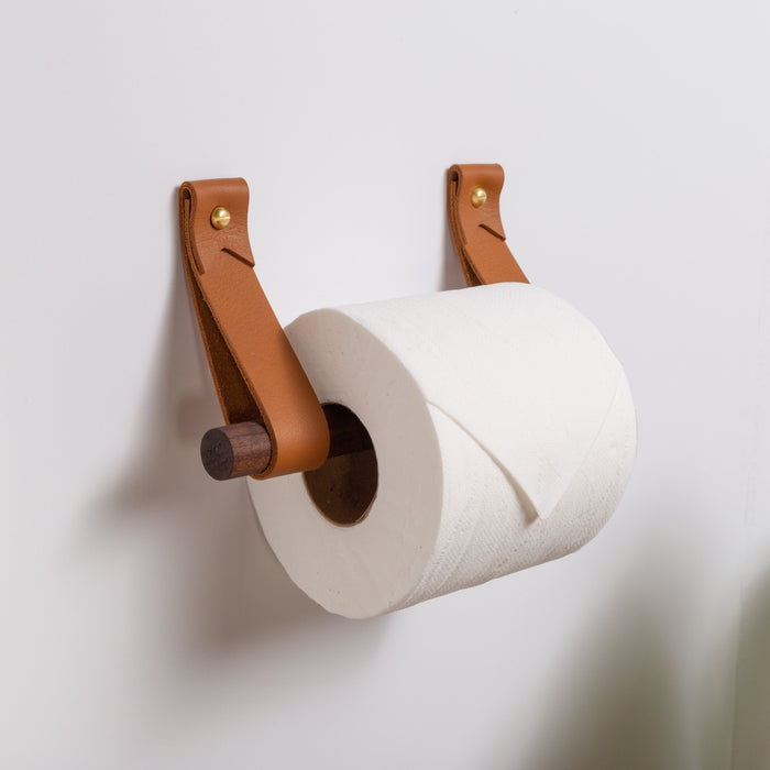 Keyaiira modern toilet paper holder crafted with nicotine-colored vegetable-tanned leather straps and solid brass hardware, mounted to a bathroom wall and holding a roll on a dark walnut dowel — minimalist Scandinavian bathroom accessory made from natural materials.