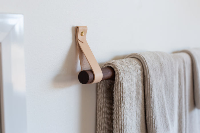 Keyaiira hanging towel rack featuring natural veg-tan leather straps with brass hardware and a dark walnut dowel, shown holding a textured beige bath towel — handcrafted modern bathroom décor designed for Scandinavian, minimalist, and organic interiors.