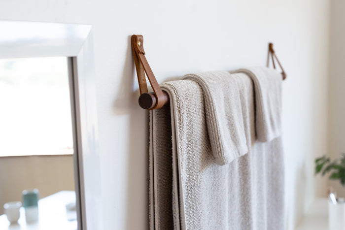 Keyaiira hanging towel rack made with nicotine-colored nicotine leather and brass hardware, holding a dark walnut dowel with a textured beige bath towel handcrafted modern bathroom storage for minimalist, Scandinavian, and organic home décor.