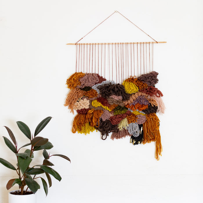 A handwoven tapestry wall hanging featuring a variety of natural fibers in brown, gray, and other colors, hung on a skinny birch wood dowel.