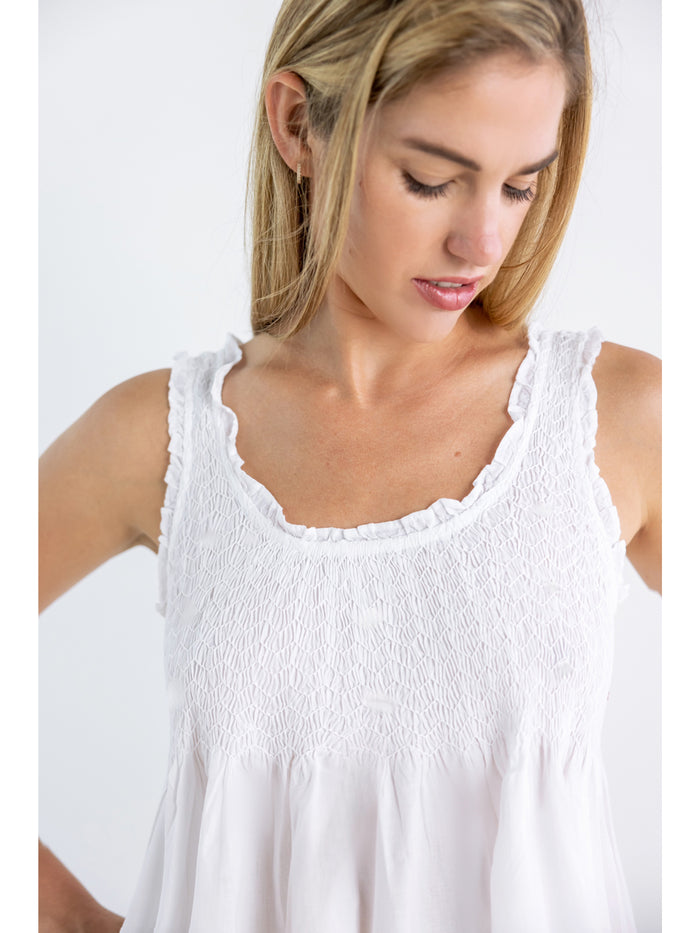 A woman wearing a white cotton nightgown with smocked detailing on the bodice and a scooped neckline with ruffle edging.