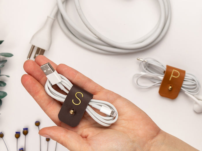 Chocolate leather cord organizer with S initial shown securing white earbuds alongside a Nicotine P cable keeper, functional Keyaiira storage for headphones, chargers and small device cords