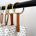 Nicotine colored leather curtain ring hanger with brass ring holding patterned white curtain on a black rod, handcrafted Keyaiira design that adds refined length and modern style to  bathrooms.