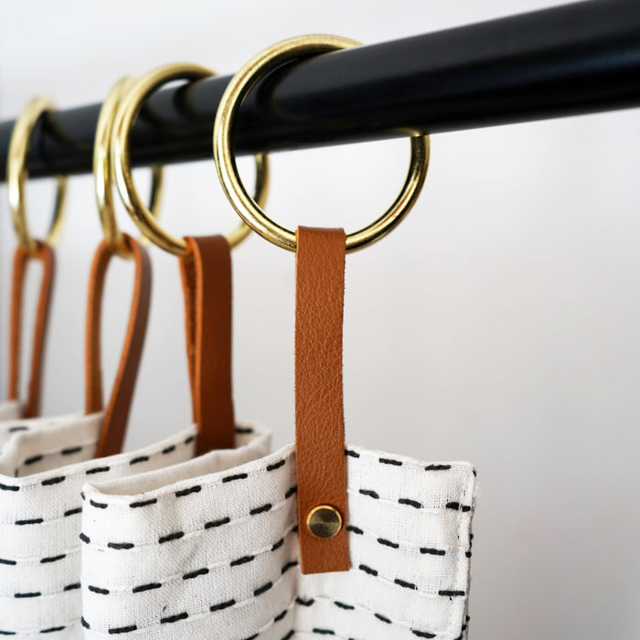 Nicotine colored leather curtain ring hanger with brass ring holding patterned white curtain on a black rod, handcrafted Keyaiira design that adds refined length and modern style to  bathrooms.
