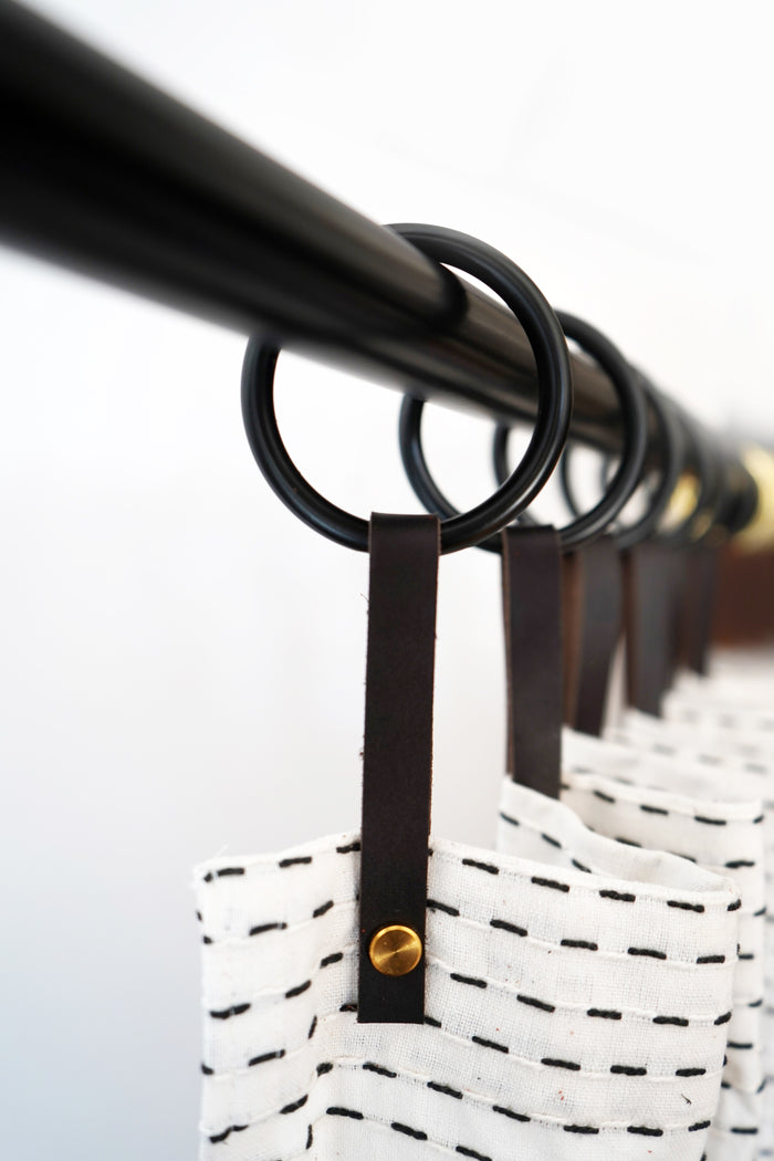 Chocolate leather shower curtain hanger with matte black ring holding a stitched white shower curtain, highlighting minimalist hardware ideal for contemporary or industrial-style bathrooms.