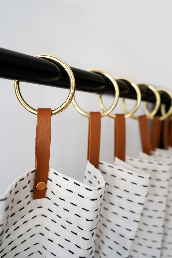 Multiple nicotine colored leather shower curtain hangers with brass rings evenly spaced along a black shower rod, adding warmth, texture, and extended drape to a minimalist shower curtain.