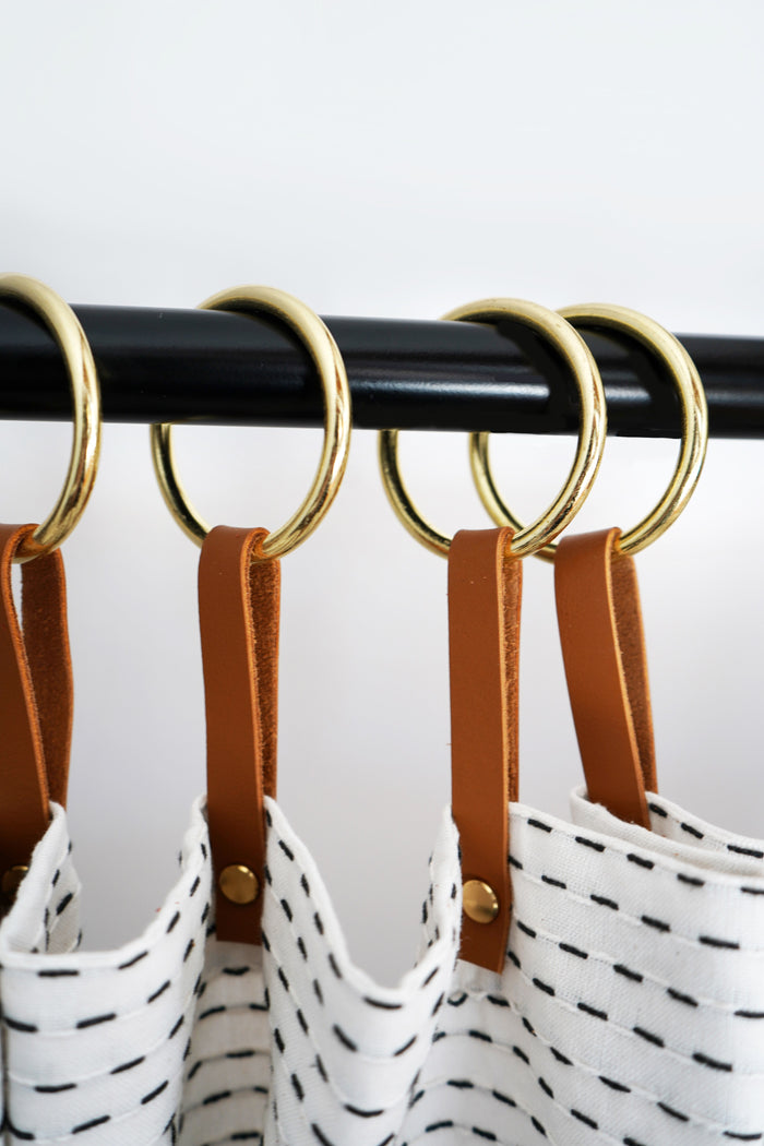Nicotine leather hangers with brass rings supporting a white patterned shower curtain, designed to enhance bathroom aesthetics while offering added curtain length and smooth sliding function.