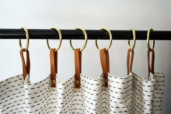 Tan leather shower curtain rings arranged on a black rod holding a stitched fabric curtain, emphasizing handcrafted hardware that blends natural materials with bathroom-friendly durability.