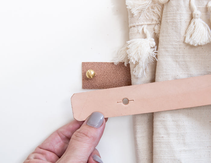 Close-up of hand installing Keyaiira natural vegetable-tanned leather wall strap with brass screw, wrapping around linen curtain — modern leather curtain tie-back detail.