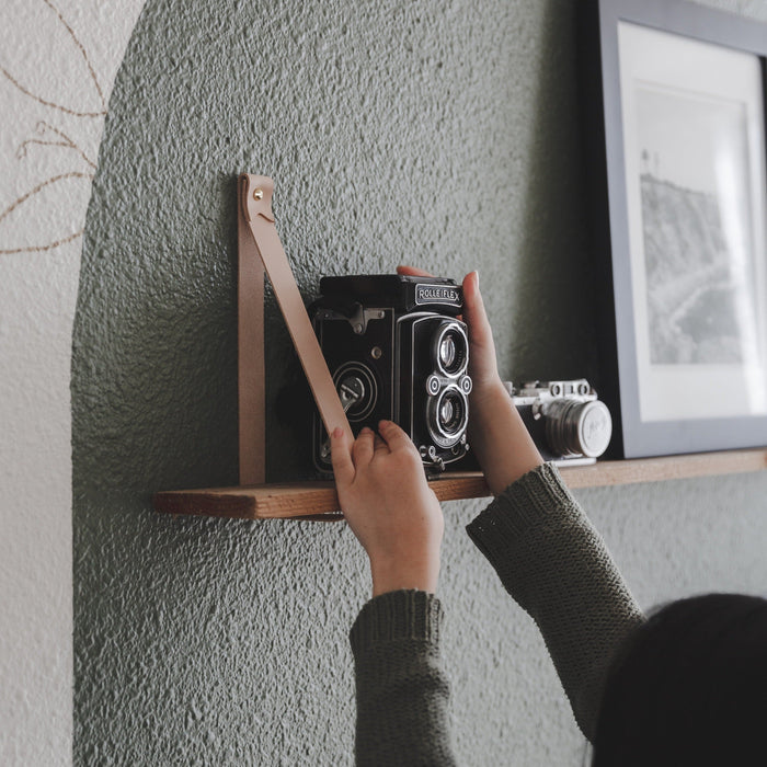 Keyaiira custom leather wall strap in standard width securing a floating wood shelf — handcrafted vegetable-tanned leather wall bracket styled with vintage camera decor in a modern minimalist interior.