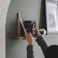 Keyaiira custom leather wall strap in standard width securing a floating wood shelf — handcrafted vegetable-tanned leather wall bracket styled with vintage camera decor in a modern minimalist interior.