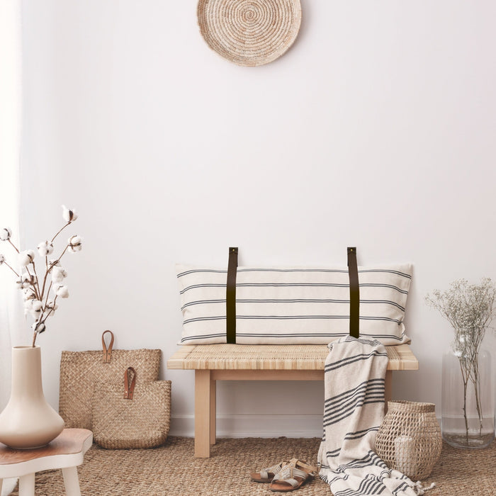Keyaiira leather wall straps shown holding a striped pillow on a woven bench — handcrafted leather loops for custom cushion or headboard mounting.