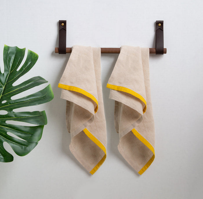 Keyaiira leather strap wall rail with wood dowel holding two neutral towels with yellow trim — minimalist leather towel bar for bathroom styling.
