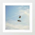 Hang'n There surreal art print — a pair of dark navy low-top trainers tied together by their laces dangling mid-air from a diagonal white jet contrail vapour trail cutting across a pale blue summer sky with soft white clouds, the classic shoe tossing shoefiti street culture act reimagined at aviation altitude, urban street art for boys bedrooms teen rooms sneakerheads and aviation lovers