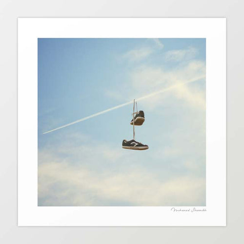 Hang'n There surreal art print — a pair of dark navy low-top trainers tied together by their laces dangling mid-air from a diagonal white jet contrail vapour trail cutting across a pale blue summer sky with soft white clouds, the classic shoe tossing shoefiti street culture act reimagined at aviation altitude, urban street art for boys bedrooms teen rooms sneakerheads and aviation lovers