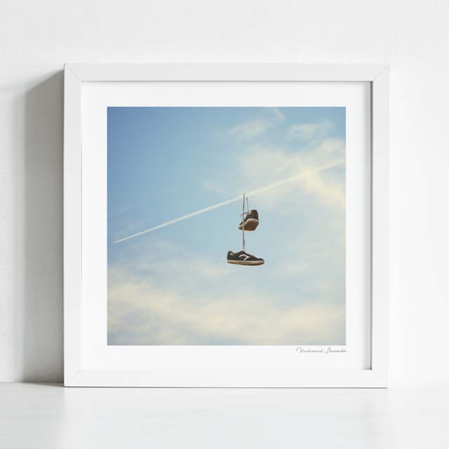 Hang'n There art print framed lifestyle view — surreal sneaker trainer shoe tossing artwork showing trainers hanging from a commercial jet contrail against a blue cloudy sky, street culture meets aviation surrealism wall art for man caves skate shops streetwear boutiques teen bedrooms and unique gift for sneaker collectors