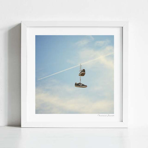 Hang'n There art print framed lifestyle view — surreal sneaker trainer shoe tossing artwork showing trainers hanging from a commercial jet contrail against a blue cloudy sky, street culture meets aviation surrealism wall art for man caves skate shops streetwear boutiques teen bedrooms and unique gift for sneaker collectors