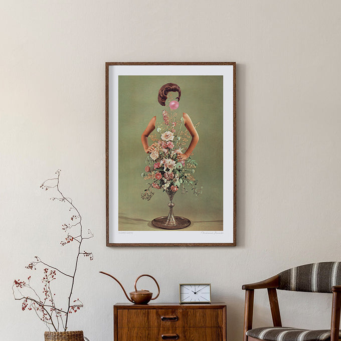 Surreal retro floral art print — a confident 1960s woman with hands on hips, blowing a pink bubblegum bubble, her torso a silver pedestal vase bursting with blush roses, cream peonies and trailing wildflowers against a sage green background, kitsch botanical fashion wall art by Vertigo Artography