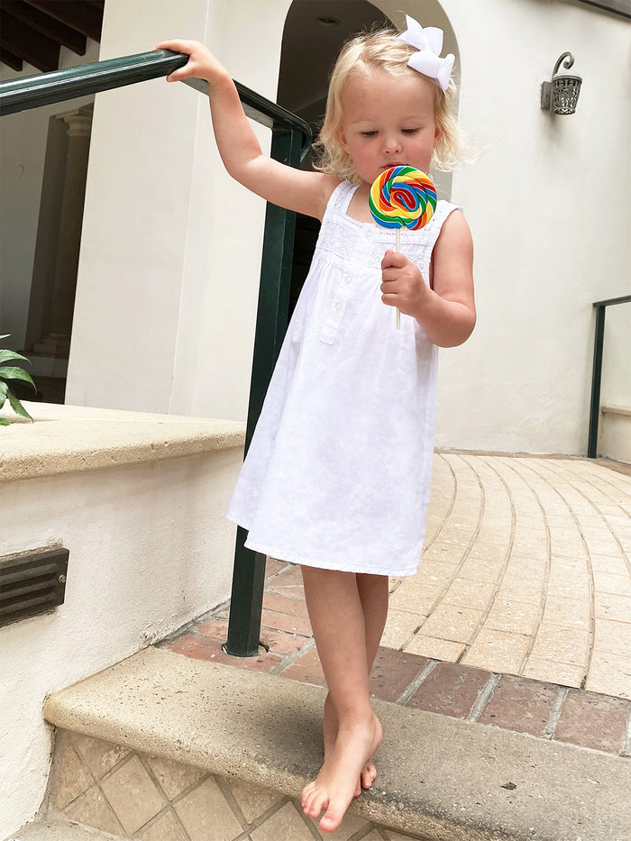A young girl wearing a white cotton dress with lace strap sleeves and front buttons, holding a colorful lollipop.