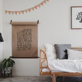 Wall-mounted kraft paper holder with walnut wood dowel and brown leather straps displayed above a bed functional modern décor for notes, artwork, menus, and lists.
