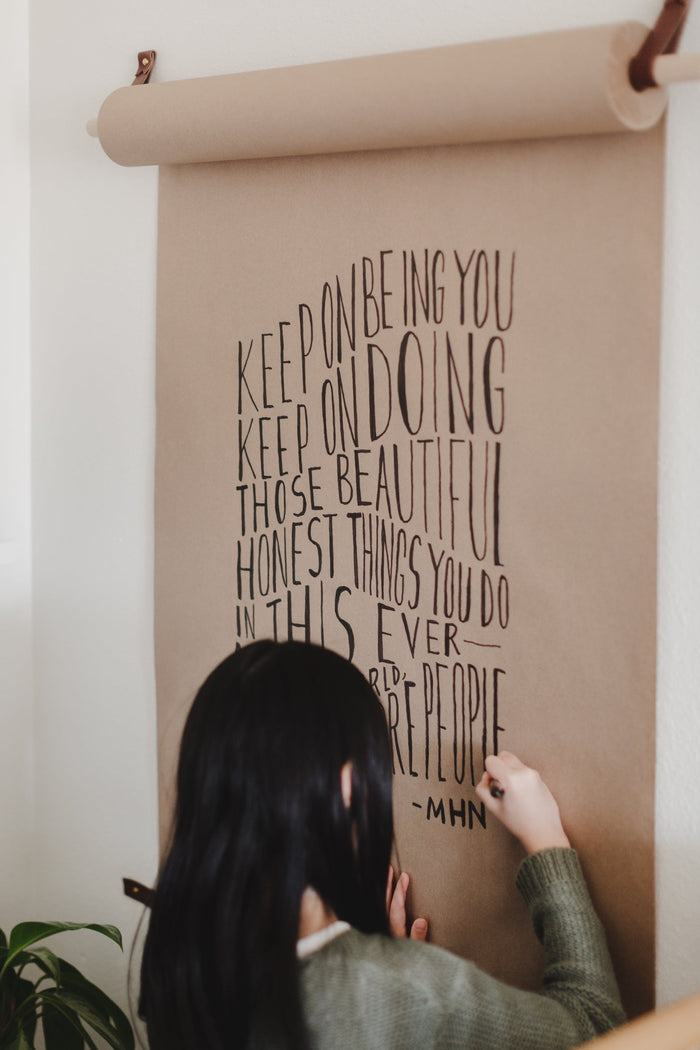 Child writing on kraft paper rolled from a minimalist wall-mounted paper holder with birch dowel and leather straps DIY message board for home or office.