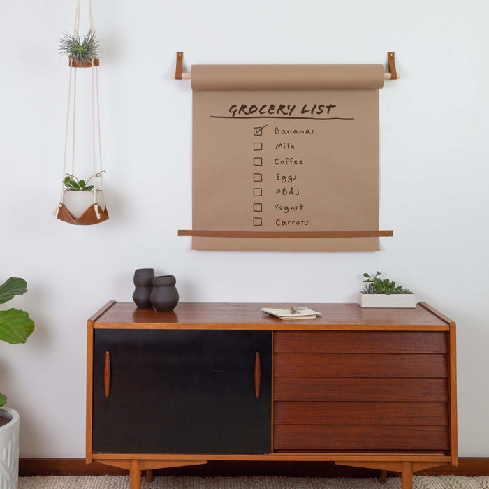 Large wall-mounted kraft paper roll holder with caramel-brown leather straps and wooden dowel, displaying handwritten inspirational quote on hanging kraft paper scroll; styled in a cozy minimalist room with white walls, interior plant in patterned pot, and a floating shelf holding small notebooks and décor.