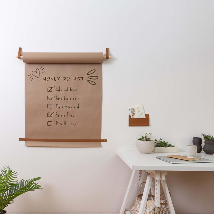 Kraft paper roll holder with tan leather straps and wooden dowel mounted on a white wall, featuring a handwritten grocery list on large kraft paper sheet; styled with modern hanging plant pot and minimalist black ceramic containers for functional kitchen or pantry organization.