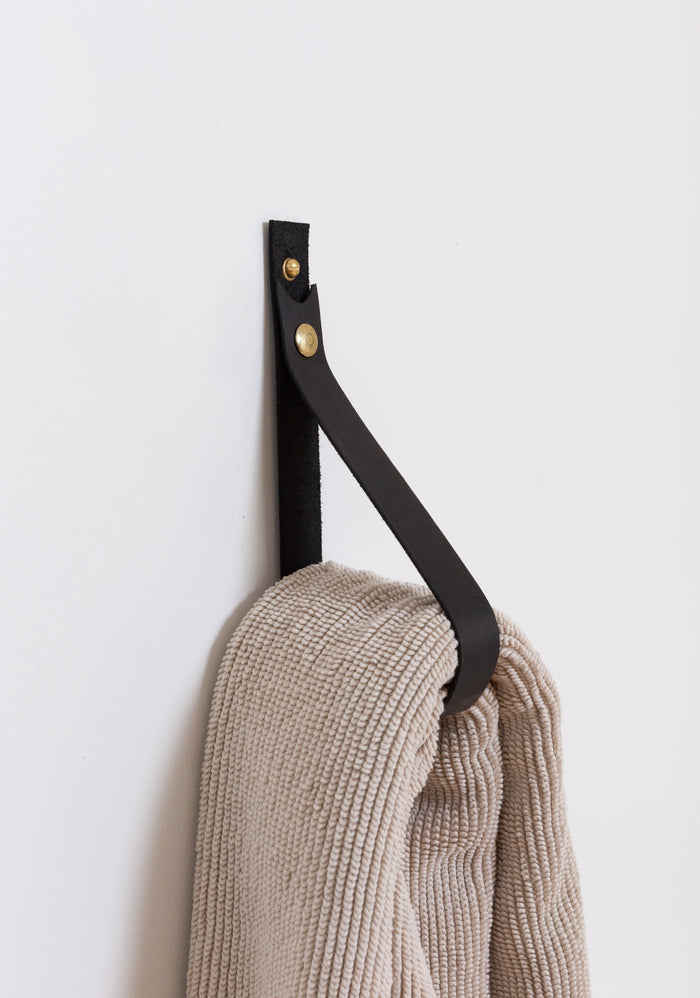 Large black leather wall strap with solid brass snap hardware mounted on a white wall and holding a neutral knit throw, showcasing Keyaiira’s handcrafted vegetable-tanned leather and minimalist hanging storage solution.