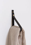 Large black leather wall strap with solid brass snap hardware mounted on a white wall and holding a neutral knit throw, showcasing Keyaiira’s handcrafted vegetable-tanned leather and minimalist hanging storage solution.