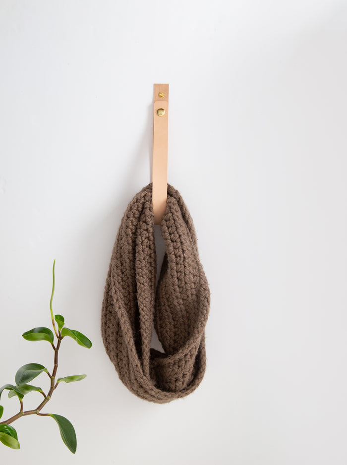 Vegetable-tan leather snap strap with brass hardware mounted on a white wall holding a chunky knit blanket, offering warm minimalist wall storage for bedrooms and living spaces.