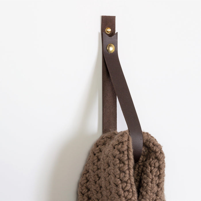 Large wall straps in chocolate leather and black leather with brass snaps holding a chunky knit throw, highlighting the contrast between deep brown and matte black vegetable-tanned hides.