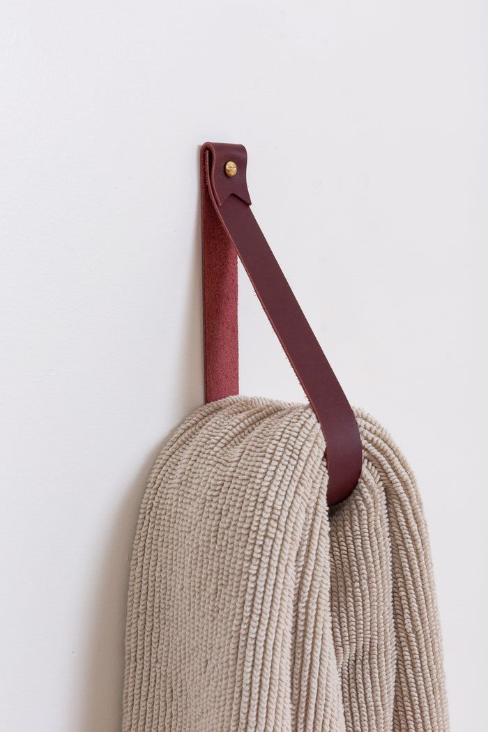 Wine-colored leather wall strap used to hang a soft beige towel on a white wall, modern handcrafted home decor accent.