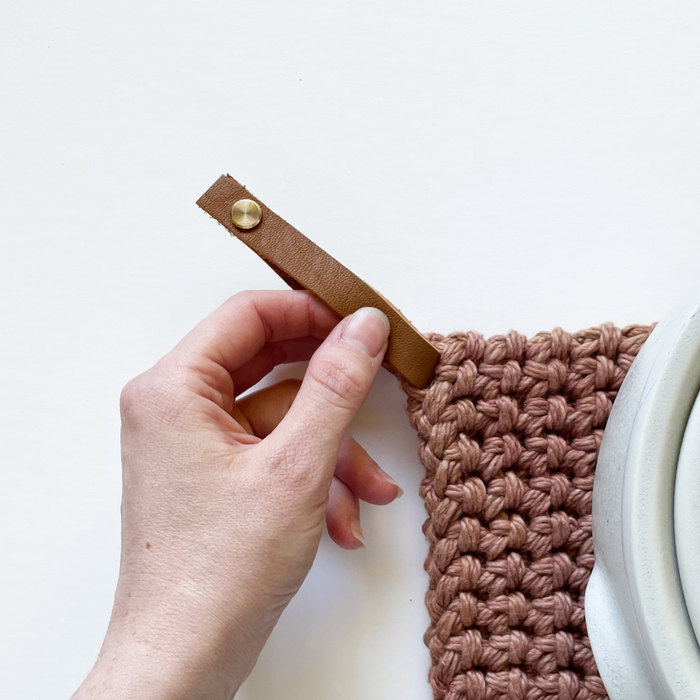 Hand attaching a Keyaiira detachable leather hanging loop to a knit potholder  handcrafted leather loop with brass hardware for kitchen organization.