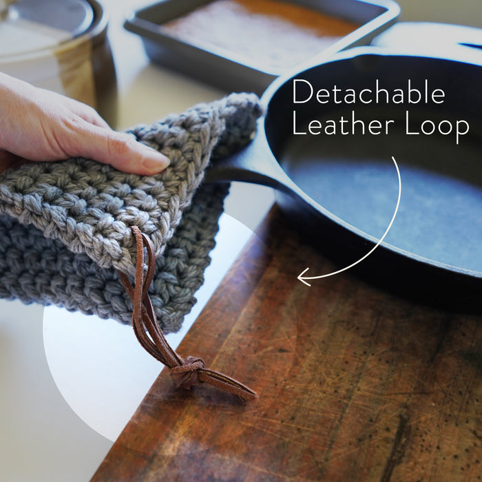 Person holding a blue-gray crocheted knit potholder with detachable leather hanging strings by Keyaiira over a wooden surface — removable leather hanging strings with brass hardware.