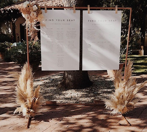 Wedding seating chart display on copper frame using Keyaiira leather signage hangers to hold minimalist printed cards with pampas grass and floral accents.