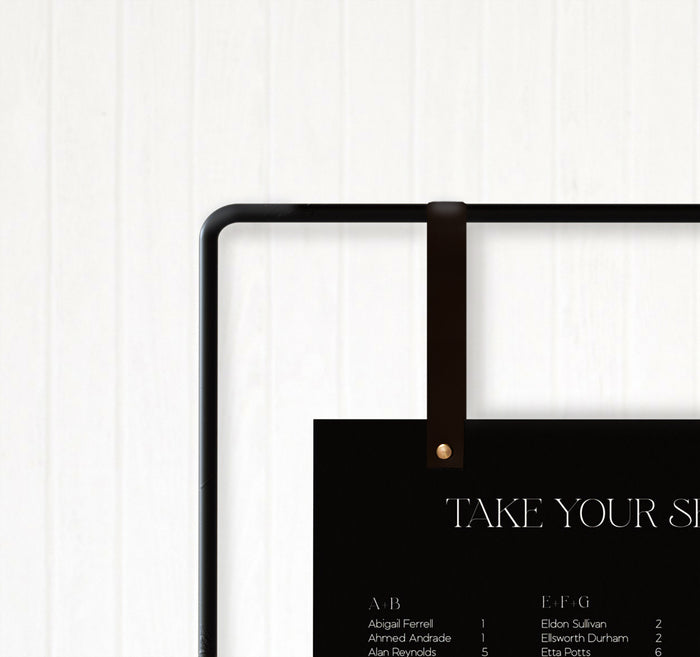 Handcrafted leather signage hanger by Keyaiira shown in black with brass hardware used to hold a ‘Take Your Seat’ wedding sign on matte metal frame.
