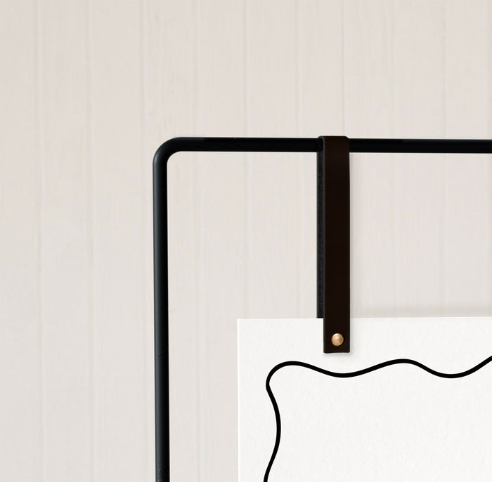 Keyaiira black leather display strap with brass hardware shown on minimalist black metal frame holding a white event sign with modern abstract edge. Handcrafted display strap for weddings, business signage, and event decor.