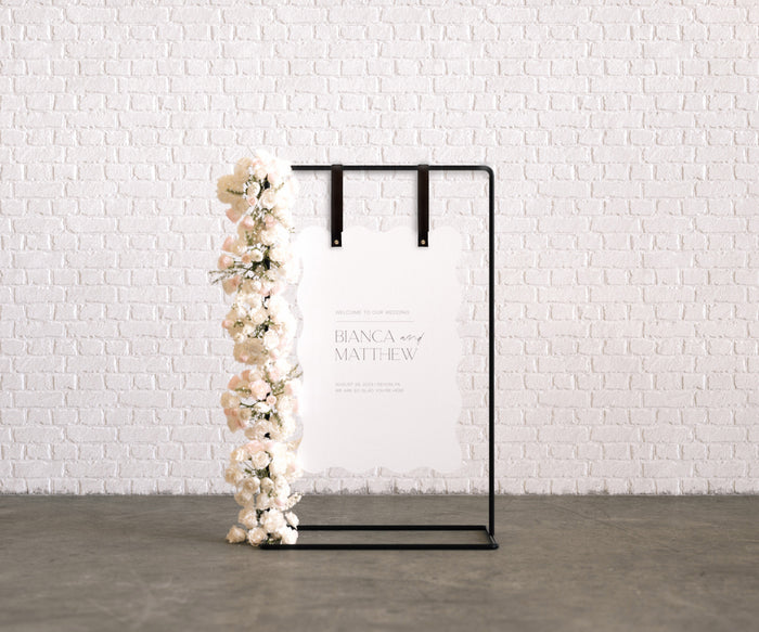 Keyaiira black leather sign straps with brass hardware used to hang a white scalloped wedding welcome sign on a minimalist black metal frame with soft floral arrangement.