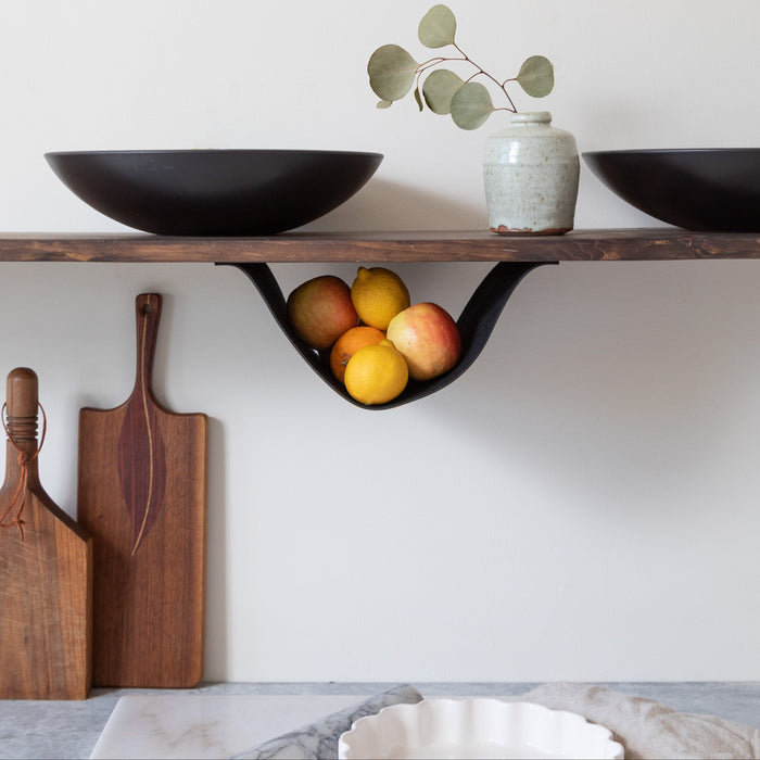 Modern black leather fruit hammock under a wooden shelf, used for under-shelf storage of apples and lemons. Minimalist kitchen organizer for open shelving, handmade leather sling by Keyaiira for stylish fruit or produce storage.