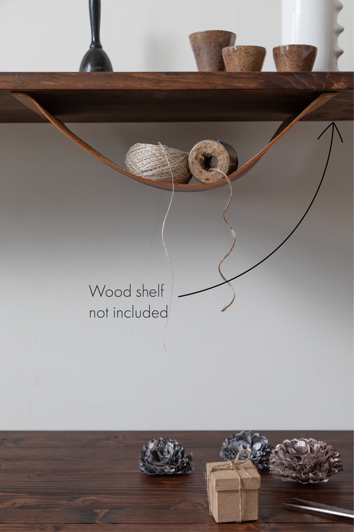 This is the Leather Sling from Keyaiira shown in use, holding yarn and twine under a wooden shelf. The handcrafted leather hammock is ideal for stylish wall or shelf storage — wood shelf not included.