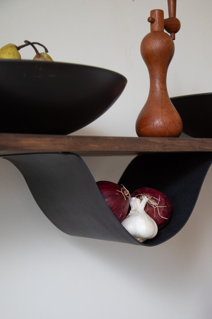 Keyaiira Leather Sling shown in use as a leather hammock holding produce under a wood shelf — handcrafted leather storage sling adds natural, modern organization to a kitchen or pantry space. Wood shelf not included.