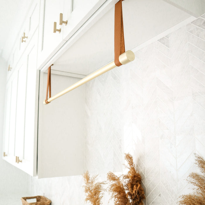 Leather suspension straps used as a curtain rod holder, mounted on a white wall with a wooden dowel rod, in a room with light decor and some plants in the background.
