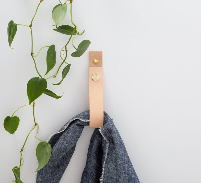 Keyaiira natural vegetable-tanned leather wall strap mounted on white wall, holding denim garment — minimalist leather wall hook with solid brass hardware for modern home organization.