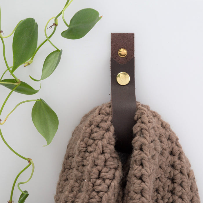 Keyaiira dark brown leather wall strap holding chunky knit throw, styled with trailing plant — modern boho leather wall hook for bedroom or living room decor.