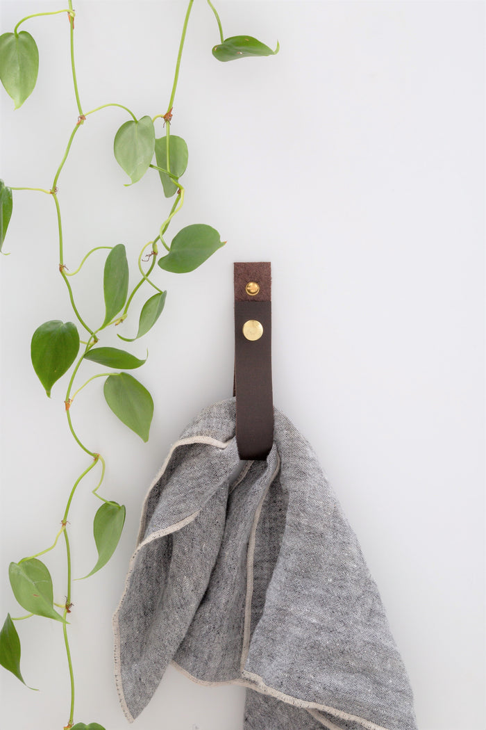 Keyaiira chocolate leather wall strap mounted on white wall with trailing greenery, securing grey towel — contemporary handcrafted leather wall organizer.