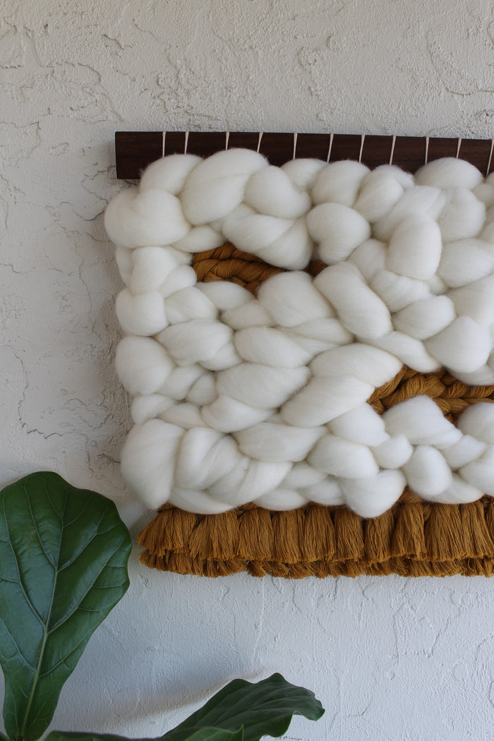 Handcrafted Keyaiira fiber wall art featuring thick natural white wool loops with a carved center detail and soft brown fringe, styled on a neutral wall with plant