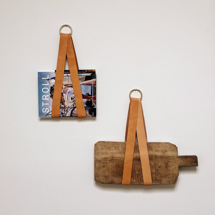 Keyaiira leather wall display holders used to hang a magazine and wooden cutting board on a white wall, showcasing artisan vegetable-tanned leather straps with brass rings for modern decorative storage.